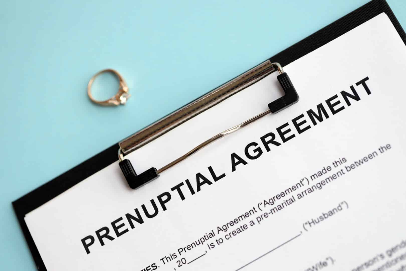 A close-up of a prenuptial agreement form on a black clipboard with an engagement ring laying on a blue counter above it.