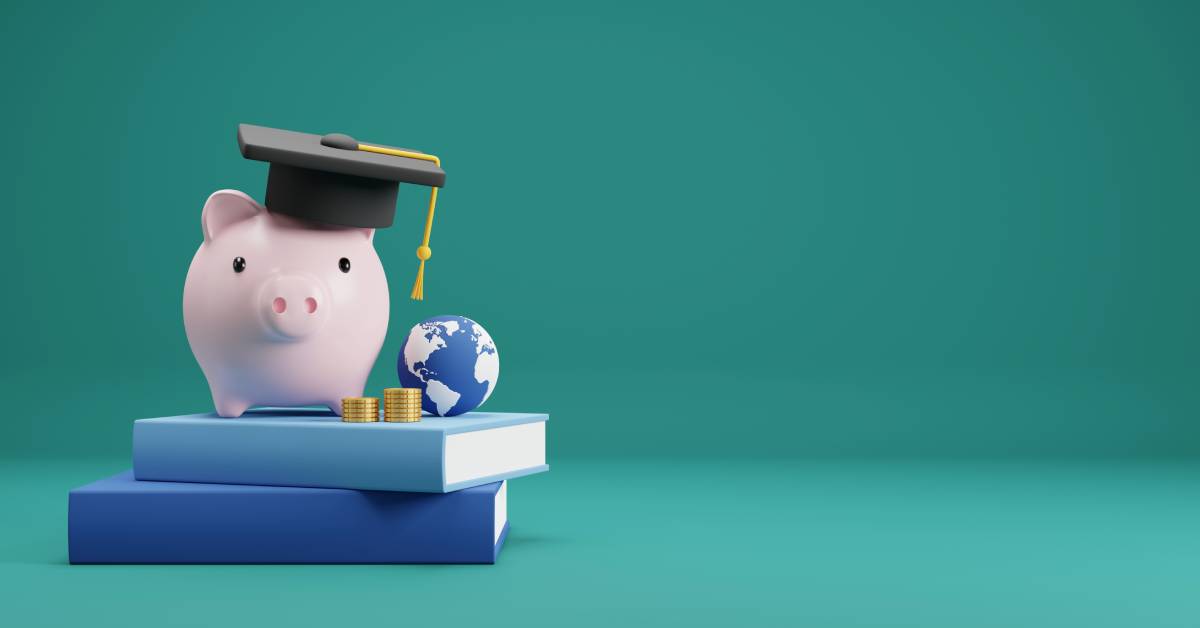 A pig figurine with a graduation cap on resting on top of two books with a globe and stacks of coins next to it.