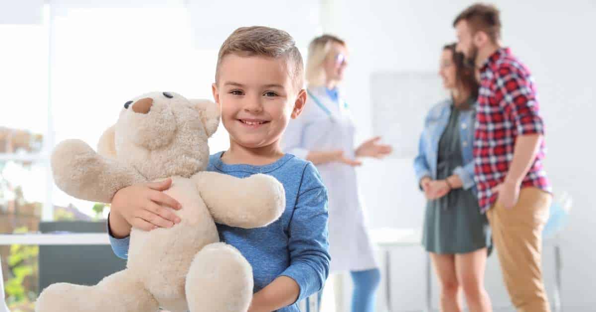 A smiling little boy holding a teddy bear while his parents discuss with a doctor in the background.