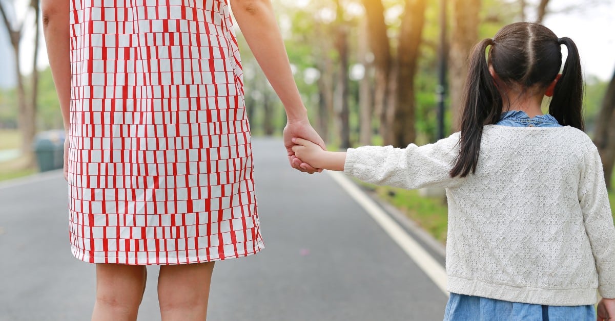 A close-up of a little girl holding a woman's hand. You can see a road, trees, and the sunlight in the background.