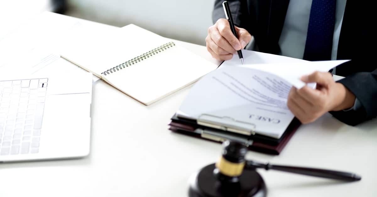 A person in a suit is looking at a clipboard and writing something in a notebook. A laptop is open on the table.