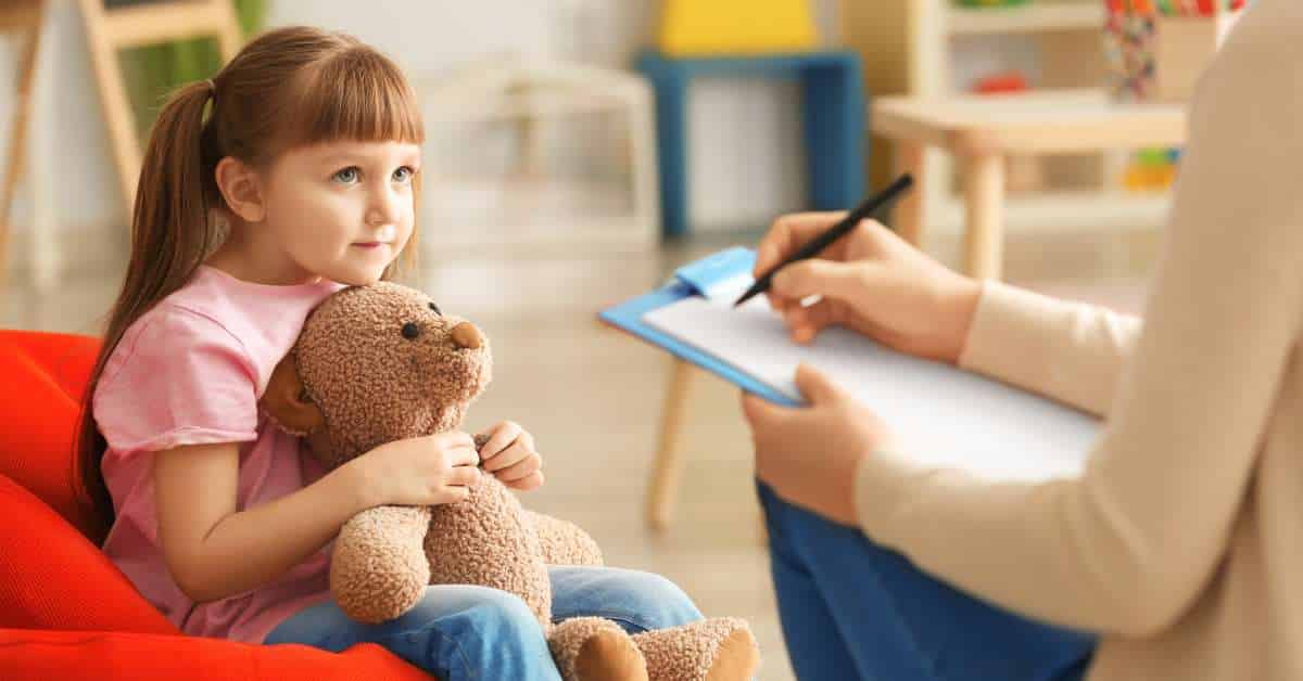 A little girl is holding a teddy bear in her lap and looking toward someone sitting in front of her with a clipboard.