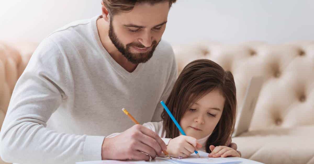 A father sits with his daughter at a small table. They are coloring on paper together with a couch in the background.