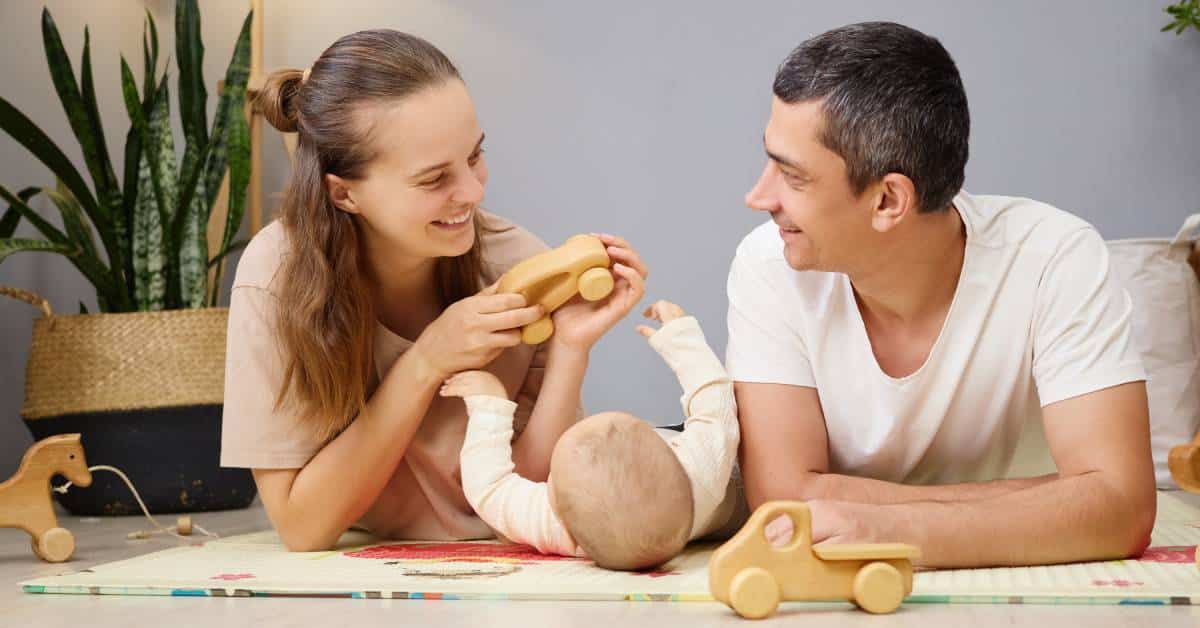 A baby lays on their back between their parents. The mom holds a wooden car, and the dad smiles at the mom.