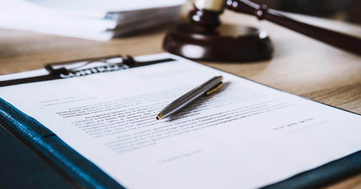 A clipboard rests on a table with a document on it. There is a pen resting on top with a gavel in the background.