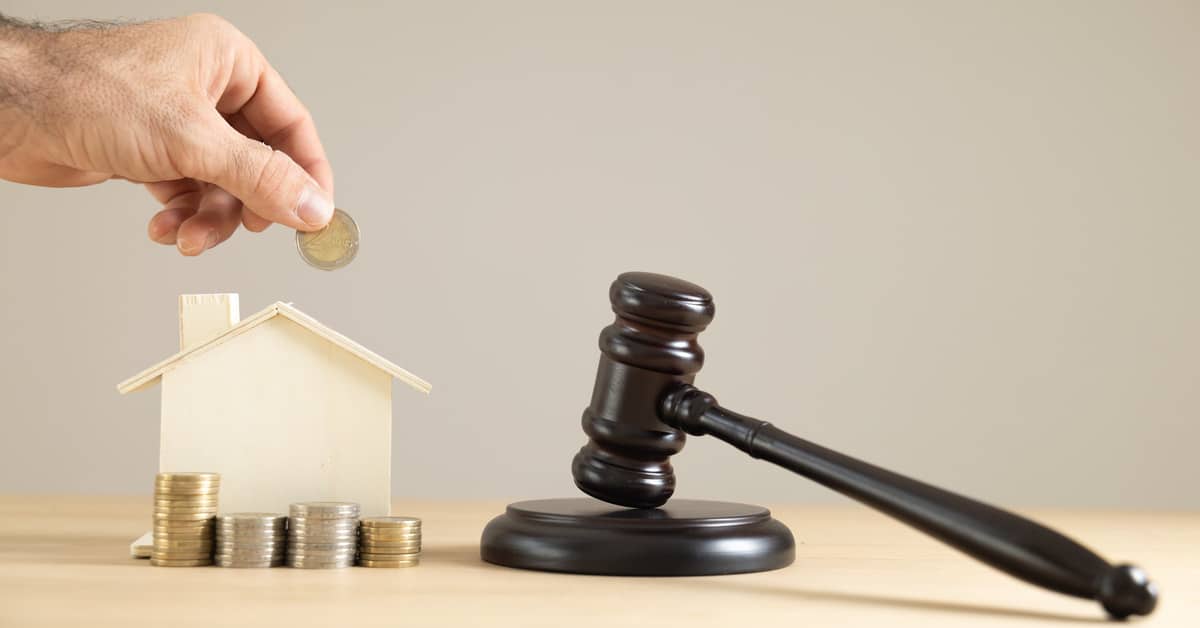 A hand holds a coin over a wooden model home with coins stacked in front of it. A judge's gavel is nearby.