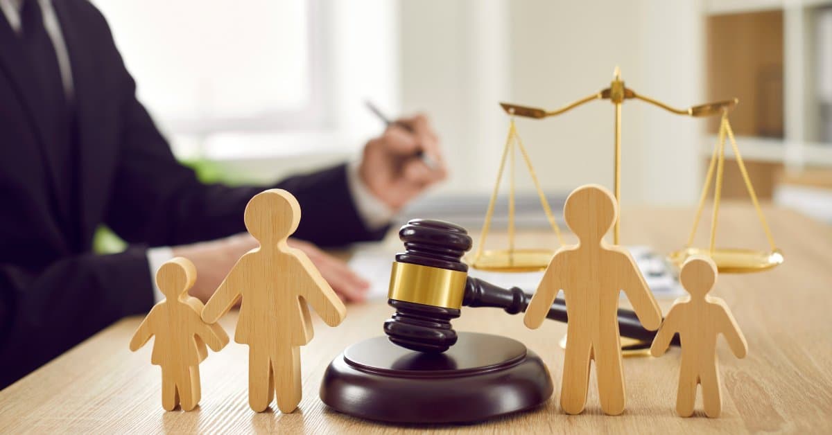 A person in a suit sits at a wooden desk. On the desk, a gavel separates wooden figures of parents and children.