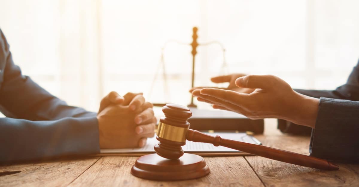 The hands of two people across from each other, with a gavel in the foreground and the scales of justice in the background.
