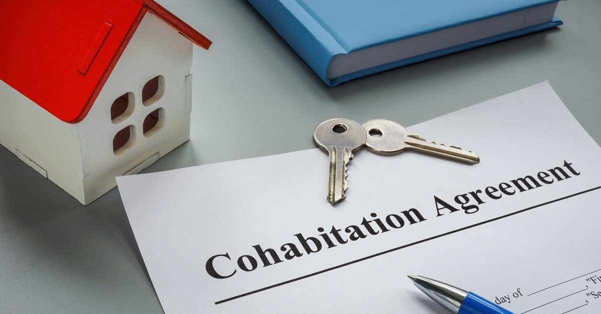 A document titled "Cohabitation Agreement" on a surface with a pen, two keys, a model house, and a book.
