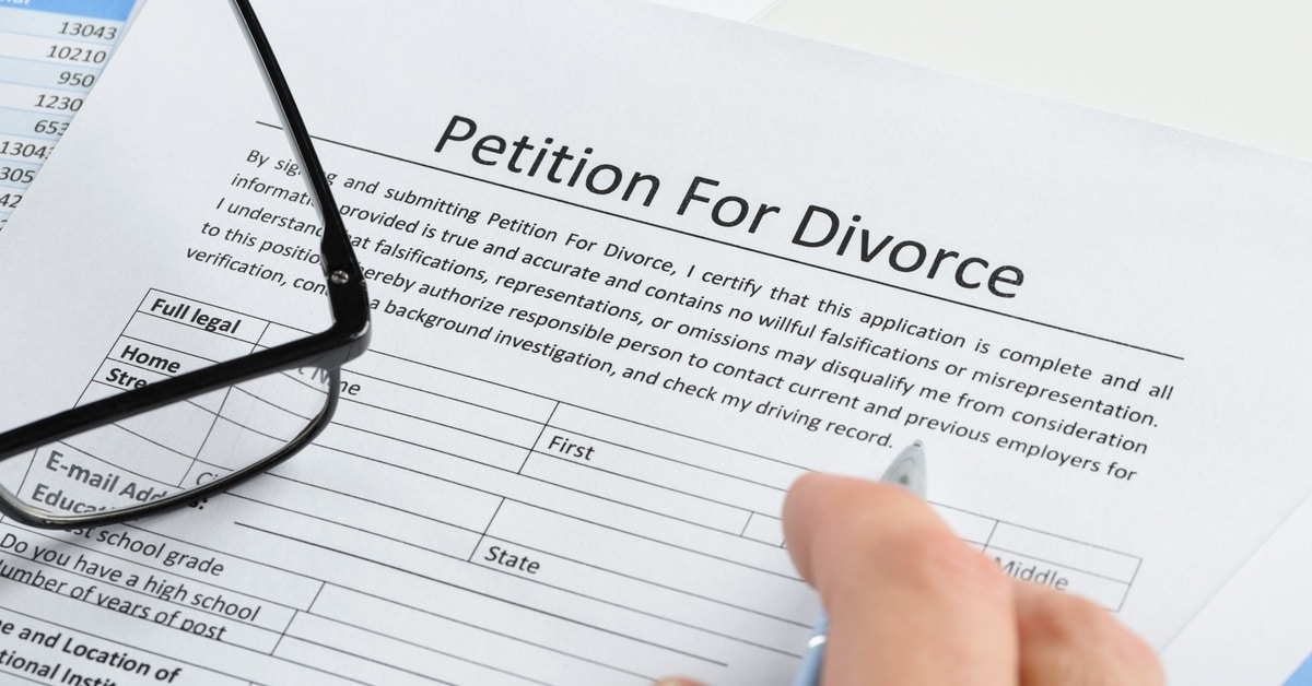 A person's hand holding a pen over a document entitled "Petition for Divorce." A pair of eyeglasses sit on the document.