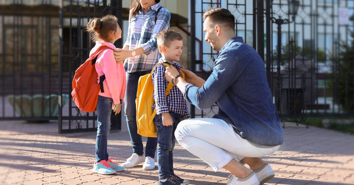 An adult woman holds a young girl wearing a backpack and an adult man kneels in front of a young boy wearing a backpack.