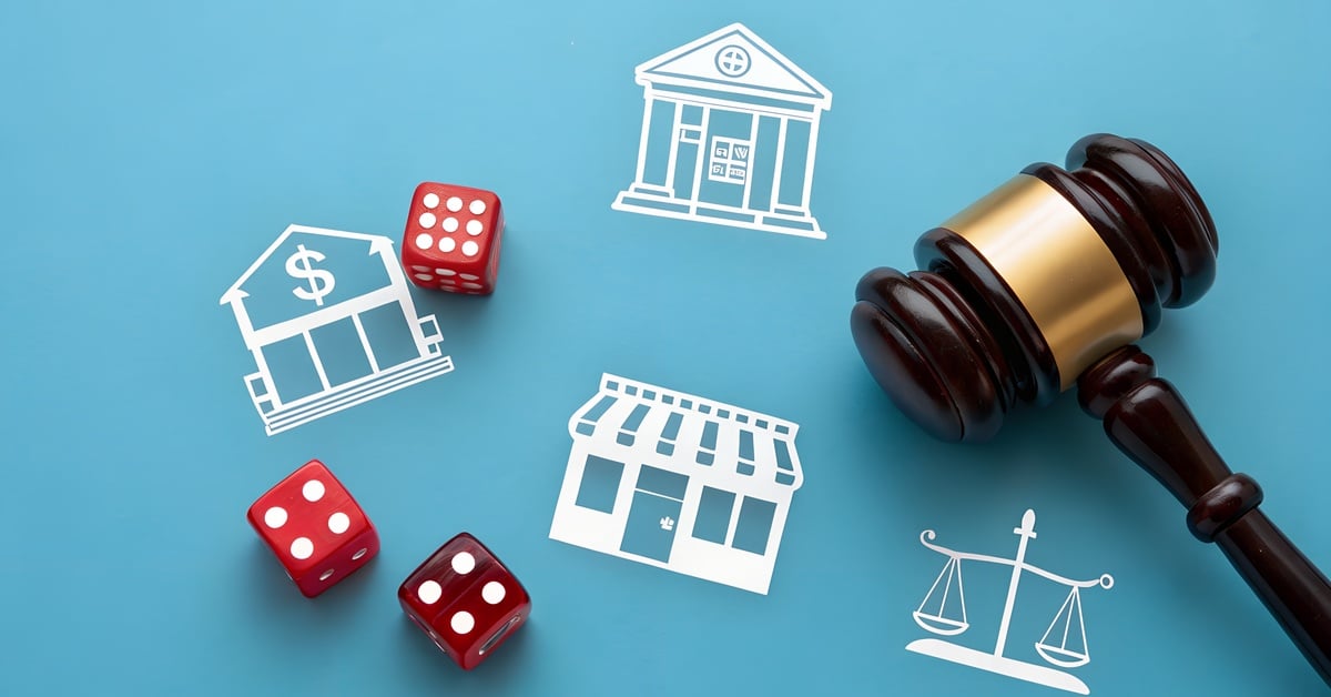 A gavel and red dice placed on a blue background with white icons related to justice, money, and a store or business on it.