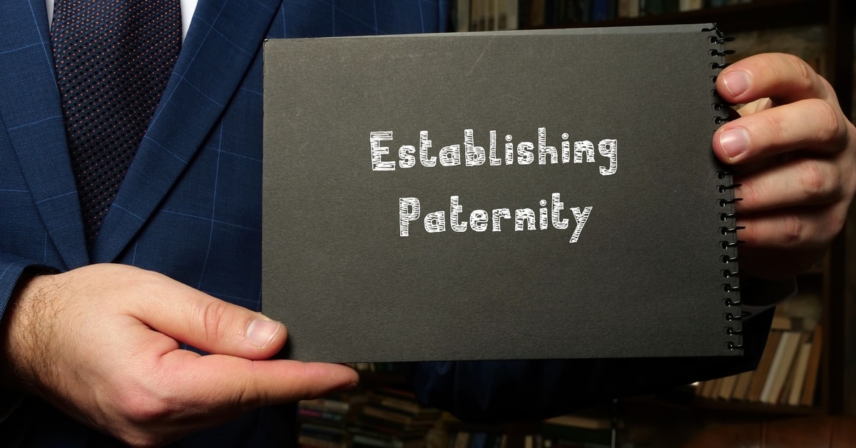 A person wearing a blue suit jacket and a dark tie holds up a notebook with the words "establishing paternity" written on it.