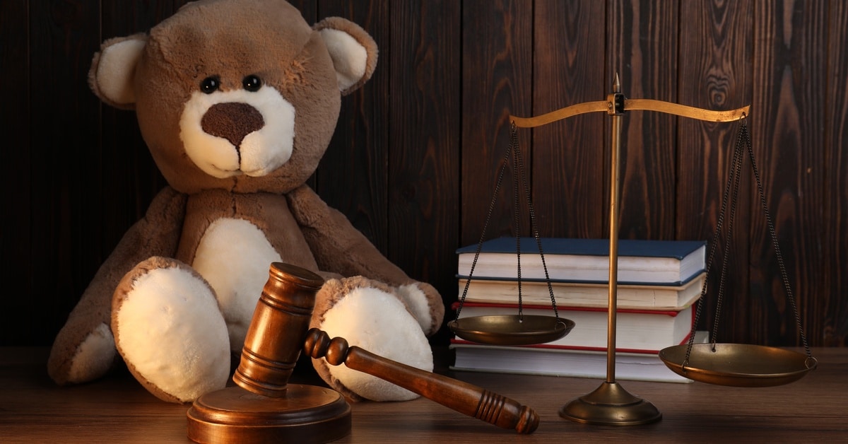 A child's teddy bear sitting on a wooden desk next to a judge's gavel, a scale, and a stack of books.