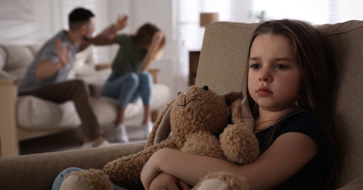 A somber little girl sitting in a sofa and holding a stuffed animal as her parents argue violently in the background.