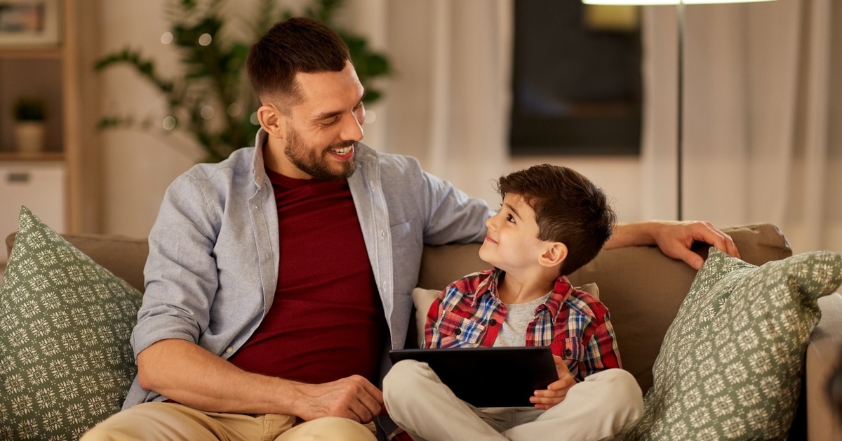 A little boy and his father sit on a living room couch at night. The boy holds a tablet and looks up lovingly at his dad.