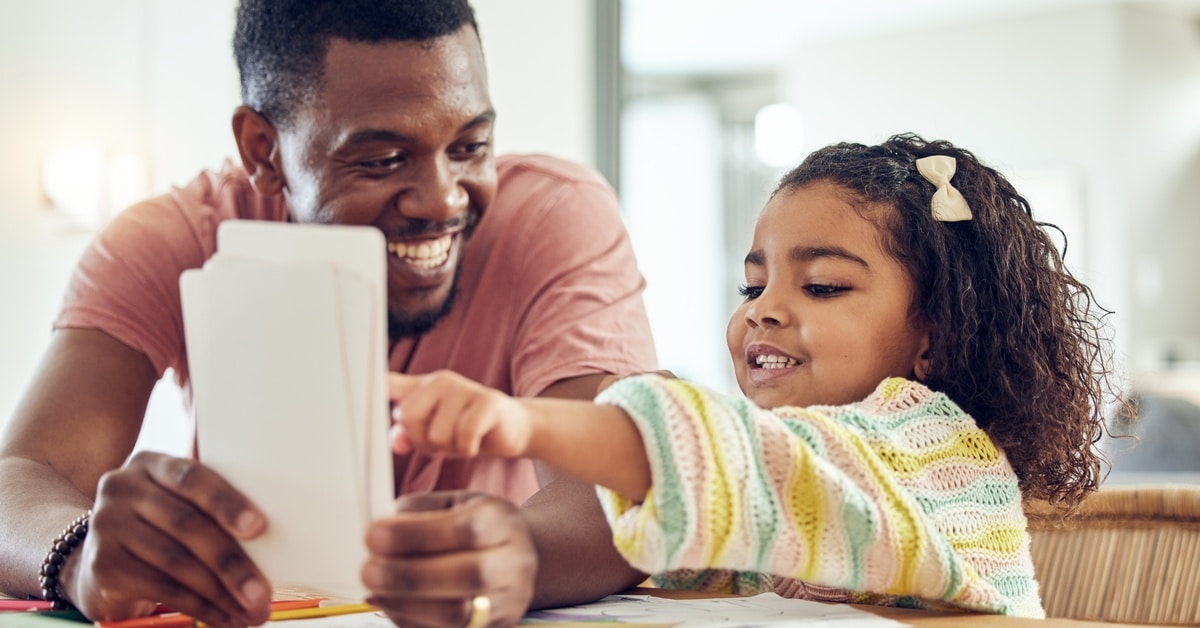 A proud, smiling father looking at his little daughter as she points to a large flashcard in his hand.