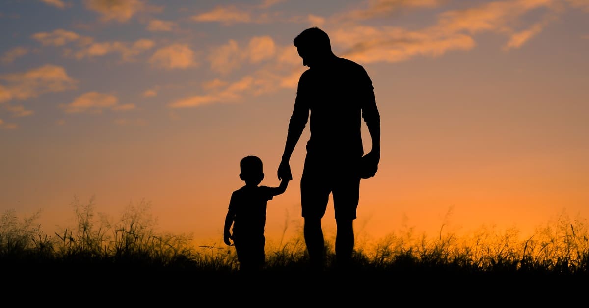 The silhouette of a father and his little son walking through a grassy field against an orange and blue sunset.