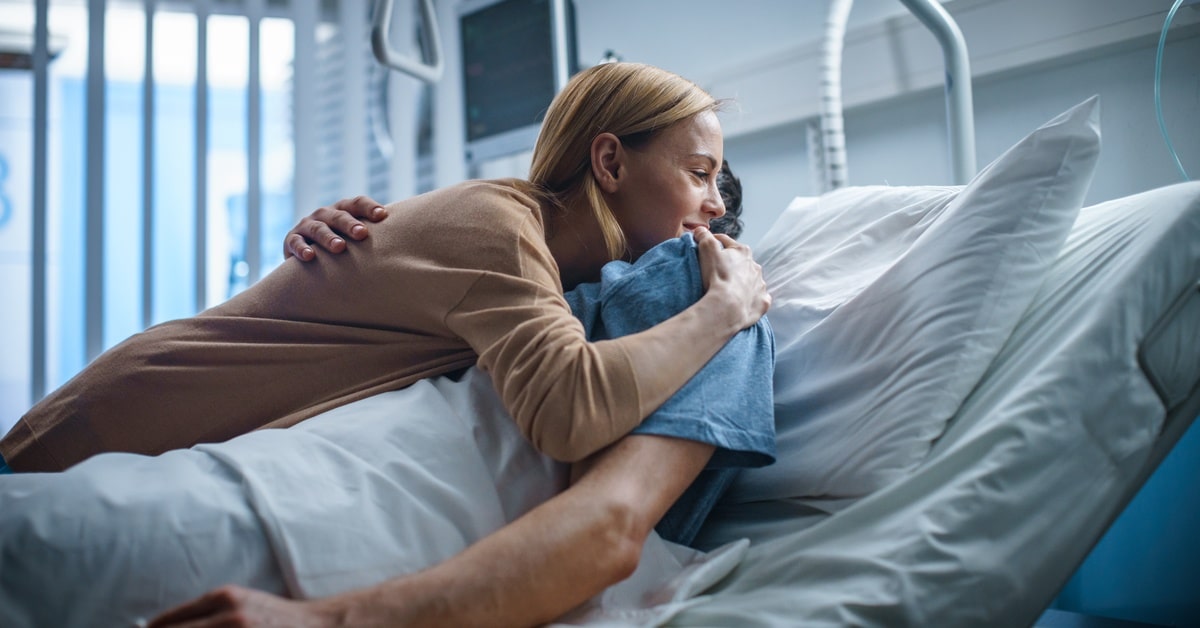 A woman in a brown shirt is leaning down to embrace her partner, who lies in a hospital bed.