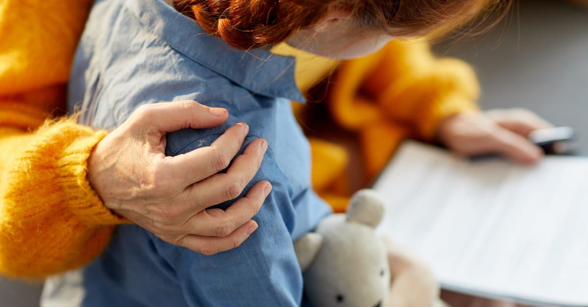 A close-up of a mother hugging her young daughter's shoulder as she reviews a document in her lap.