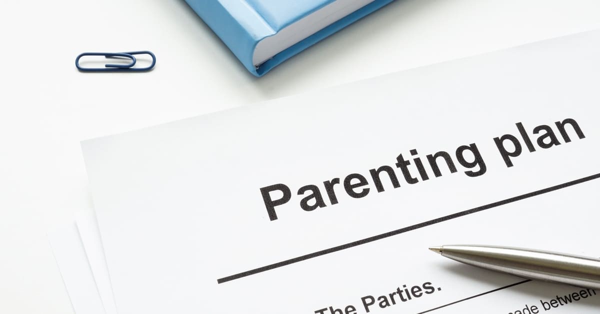 A close-up of the corner of a document titled "Parenting Plan" next to a book with a blue cover and a tiny blue paperclip.