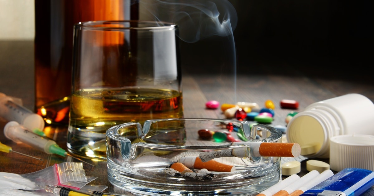 Addictive substances, including liquor, cigarettes, pills, cocaine in pouches, and drugs in needles on a wooden table.