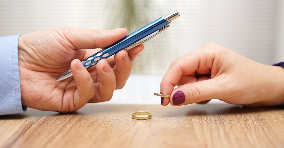 A woman's hand placing a wedding ring next to another on a table while a man's hand extends a pen to her.