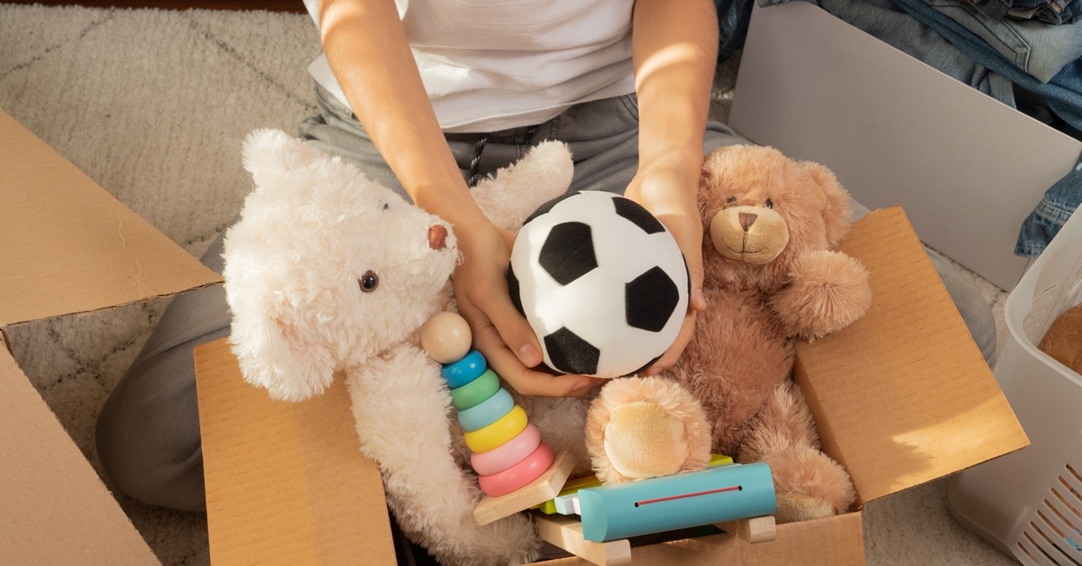 A boy holding a stuffed soccer ball over a cardboard moving box filled with teddy bears and other toys.
