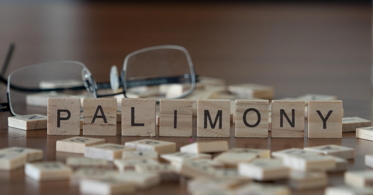 A line of wooden letter tiles spelling "PALIMONY" on a table in front of some out-of-focus eyeglasses.
