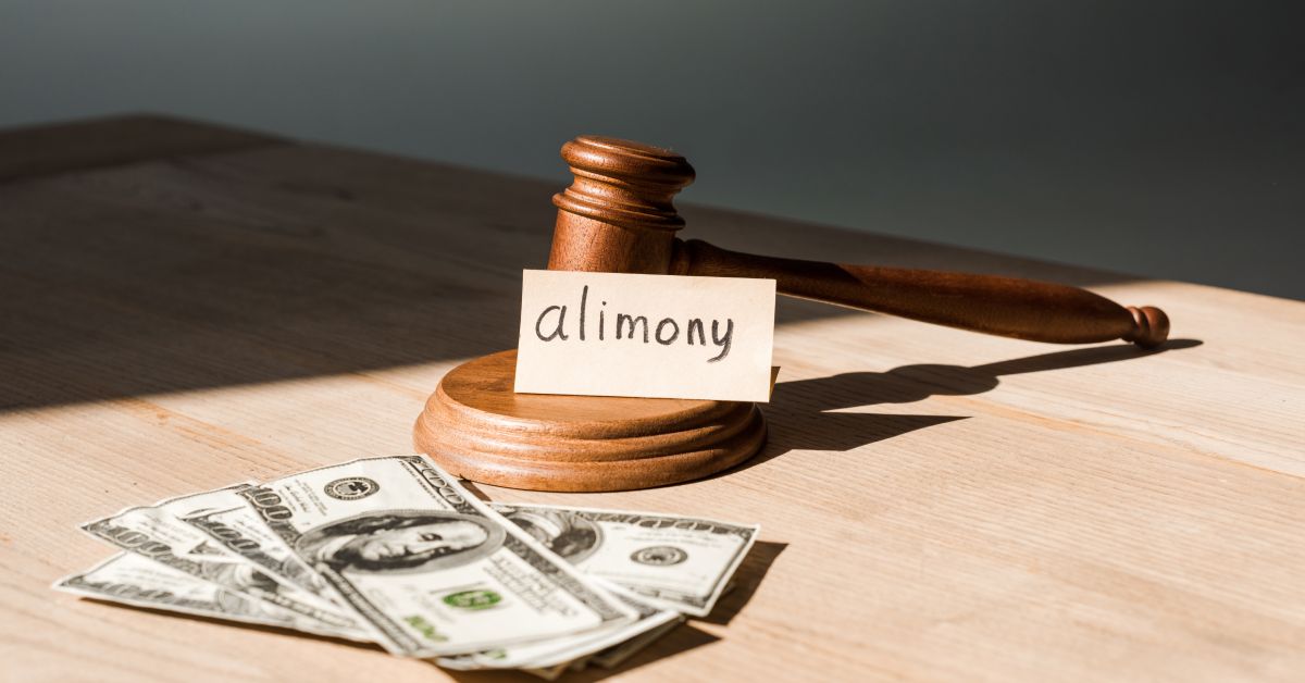 A card labeled "alimony" propped up against a wooden gavel and base next to four hundred-dollar bills on a table.