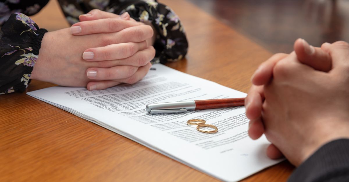 A couple sits across from each other at a table. Legal papers, a pen, and wedding rings sit between their clasped hands.