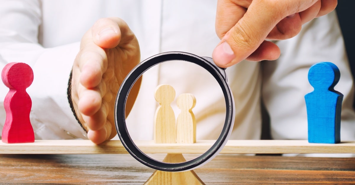Hands hold a magnifying glass over two small wooden figures between red and blue figures on a balance beam.