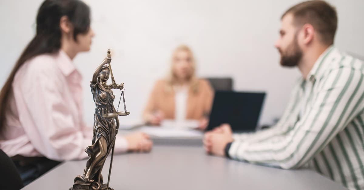 A statue of Lady Justice in front of a blurred couple discussing from either side of a table, at the end of which sits a lawyer.