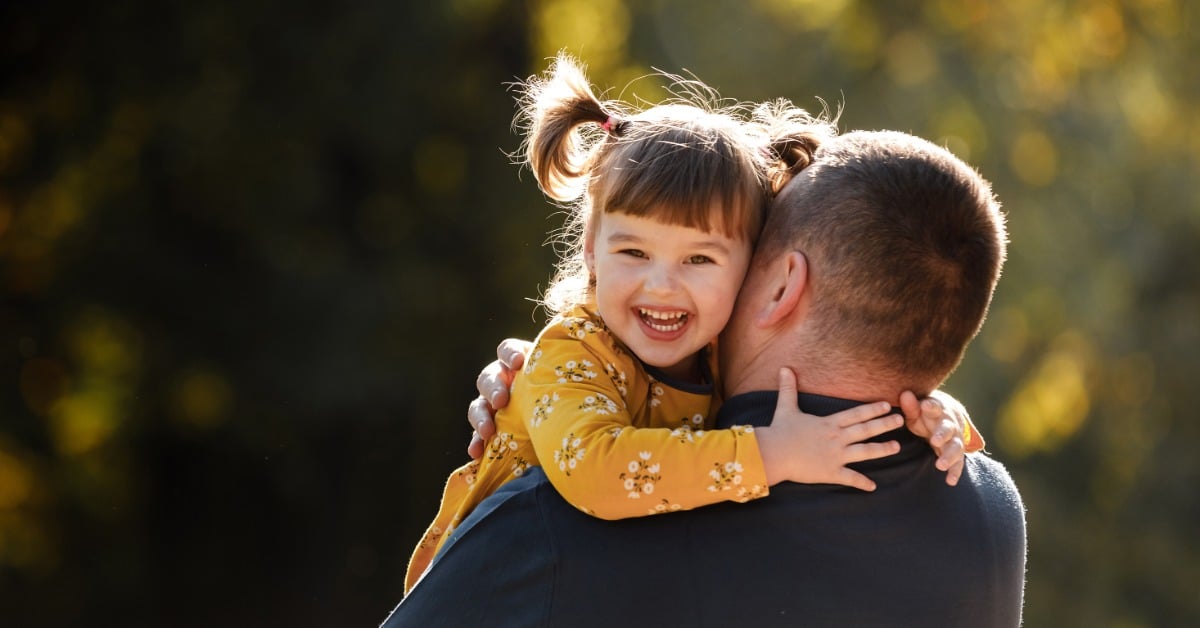 A laughing little girl in a yellow floral dress hugs her dad's shoulders as he carries her outdoors in warm sunlight.