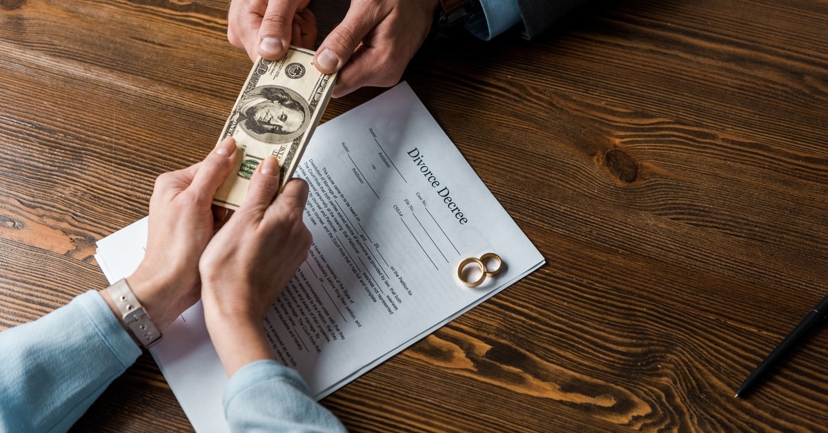 Two people exchange cash over a divorce decree on a wooden table, with wedding rings set on top of the paperwork.