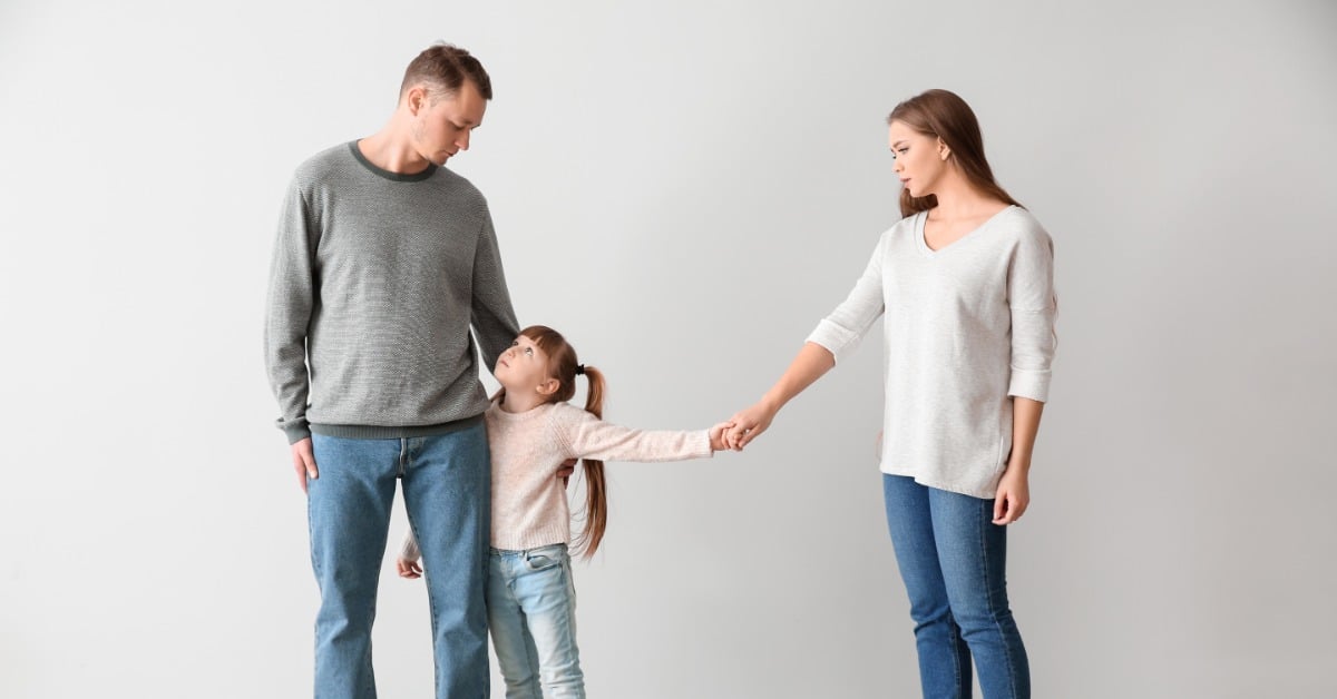 A young girl stands between her separated parents. She hugs her dad's leg and looks up at him and holds her mom's hand from afar.