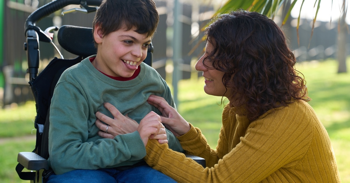 A little boy sits in a wheelchair outside with the sun shining. A woman crouches next to the boy and smiles at him.