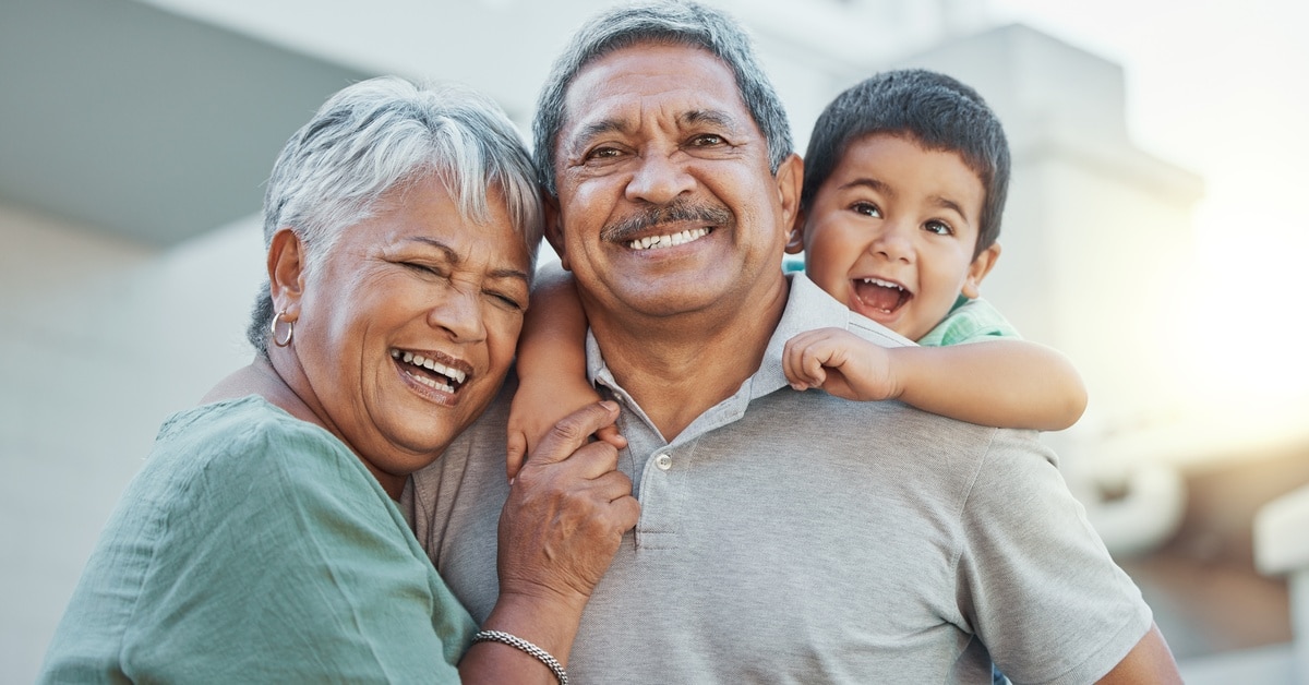 A woman with gray hair leans on an older man next to her. A child smiles as the man holds him on his back.