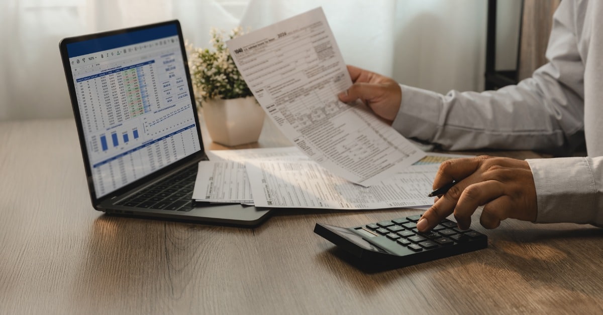 A professional reviews financial statements beside a laptop while using a calculator at a wooden desk.
