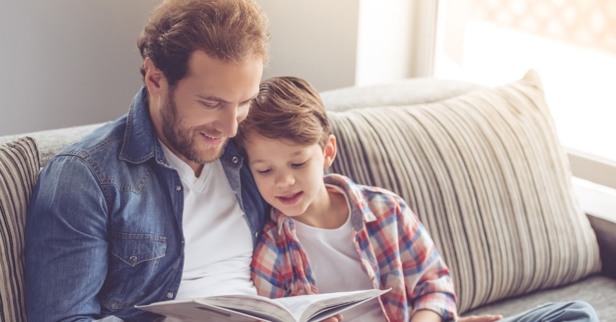 A father and his young son sit together on a couch and read an open book in a bright, sunlit living room.