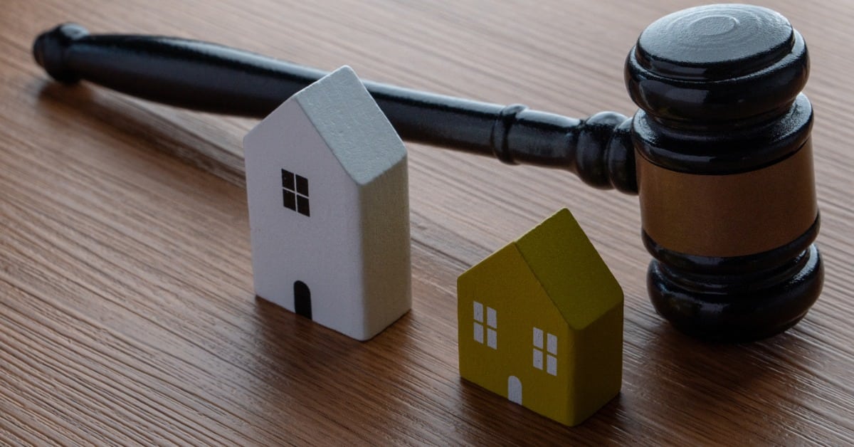 A black judge’s gavel lies beside a white house model and a yellow house model on a brown wood-grain table.