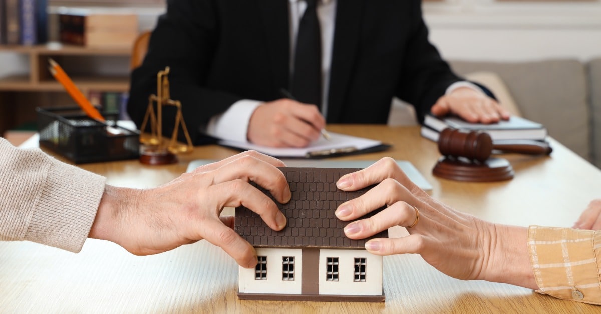 A couple holds opposite sides of a model house across a table while a lawyer sits behind a desk with a gavel.