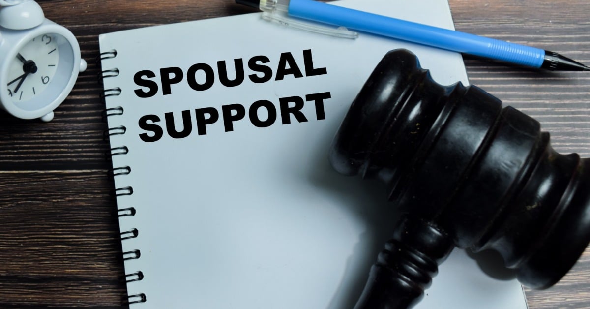 A black gavel rests on a notebook labeled "SPOUSAL SUPPORT" beside a blue pen and a white alarm clock on a wooden table.