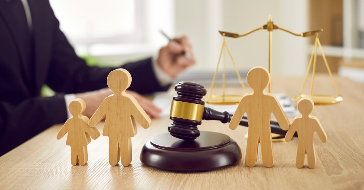 A judge’s gavel sits between wooden family figures as a lawyer writes beside the scales of justice nearby.