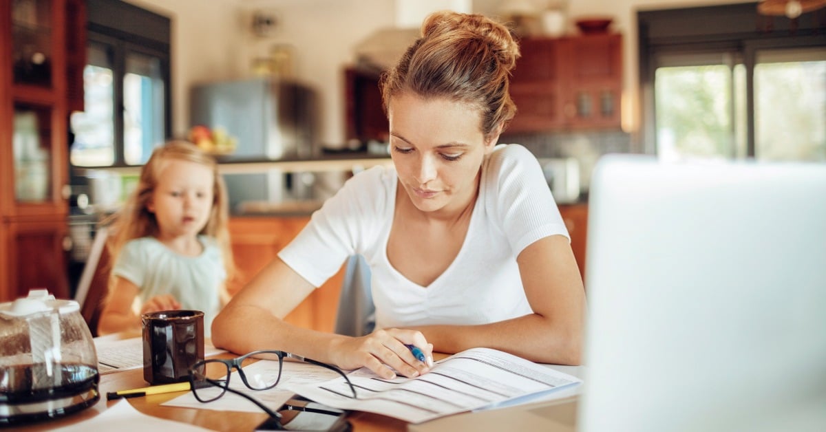 A woman sits at a kitchen table with a laptop open and paperwork in front of her. A little girl stands behind her.