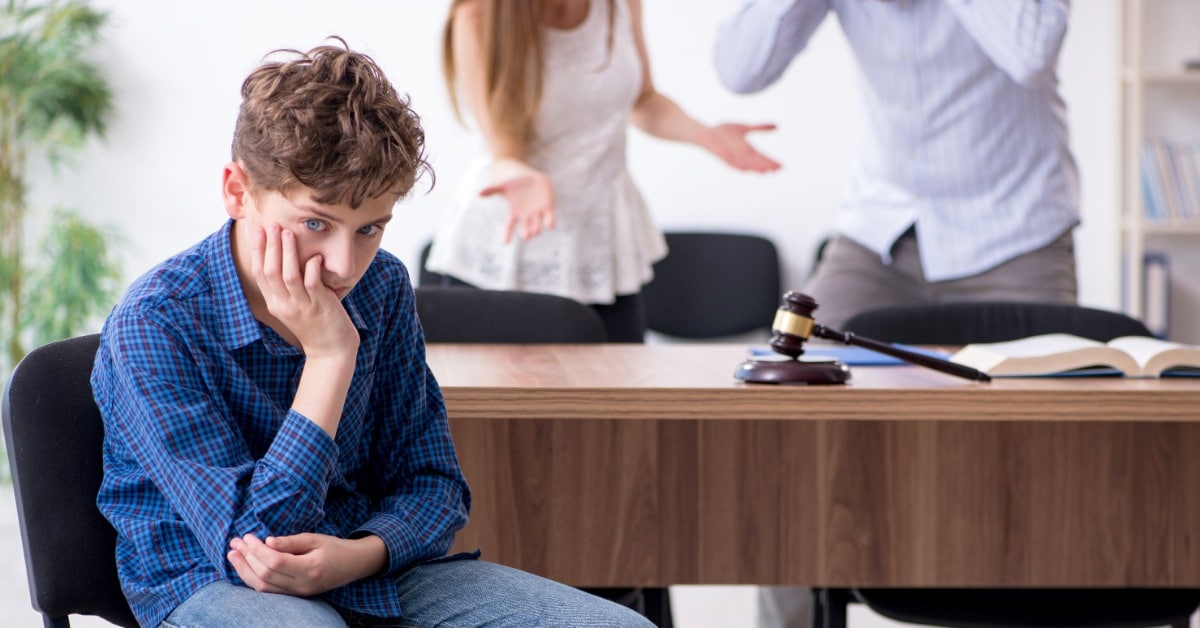 A distressed boy sits in front of a desk with a gavel on it. A couple, blurred in the background, appear to be arguing.
